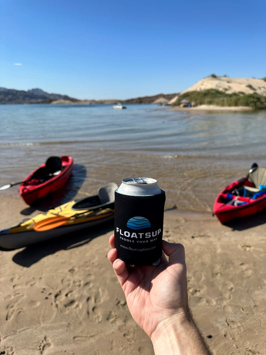 A hand holding up a can with a Floatsup coozie on it with Kayaks and a lake in the background. 